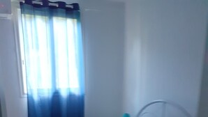 A small, single-bed room with a metal frame bed, a bedside table with a lamp, and a window with curtains. - House terrace beach riviere (pointe noire)