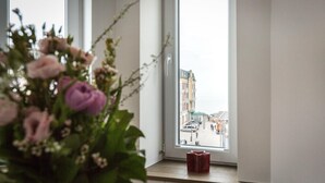 A window with a view of a street and buildings, a vase with flowers on the sill, and a red object on the windowsill. - Art your Life - Residence, Whg. 11 Superdet - Art your Life - Residence Seeburg (Sylt)