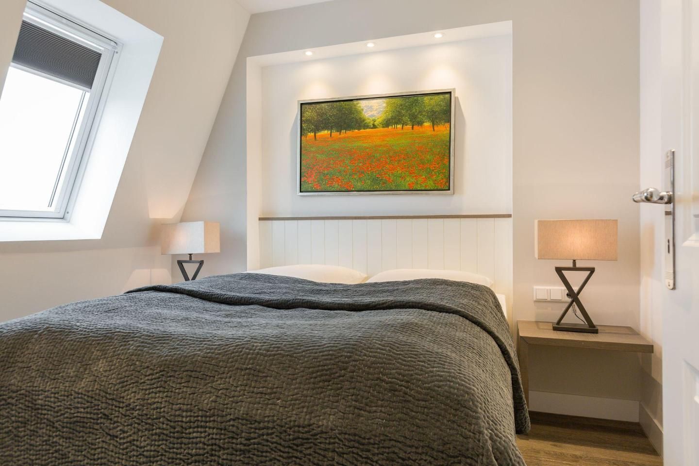 A bedroom with a bed, a painting of a field, a window, and two bedside tables with lamps.