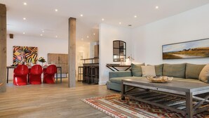 A modern living room with a sofa, a coffee table, and a dining area with red chairs. - Art your Life - Residence, Whg. 11 Superdet - Art your Life - Residence Seeburg (Sylt)