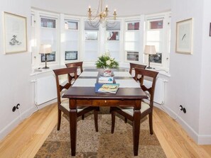 A dining room with a wooden table, four chairs, a chandelier, and a window with blinds. - Villa Maybach, Whg. 1 Albert - Villa Maybach (Sylt)