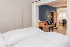 A bedroom with two beds, a sofa, and a dining area visible through an open doorway.