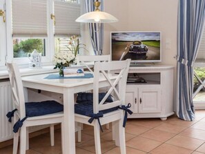 Ein Essbereich mit weißem Tisch und Stühlen, einem Fernseher und einem Fenster mit Vorhängen.