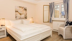 Un dormitorio con cama blanca, mesitas de noche, una silla de mimbre, una ventana con cortinas y un espejo adosado a la pared.