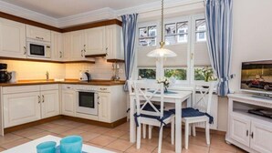 Una cocina con armarios blancos, una mesa de comedor con sillas, una lámpara colgante y un televisor sobre un mueble.