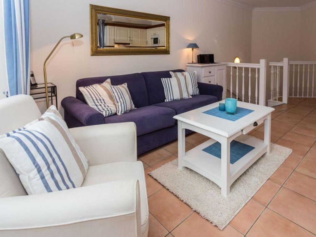 A living room with a purple sofa, white armchair, and a white coffee table. There is a mirror on the wall, a lamp, and a small cabinet.