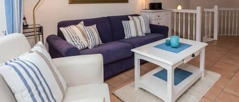 A living room with a purple sofa, white armchair, and a white coffee table. There is a mirror on the wall, a lamp, and a small cabinet.