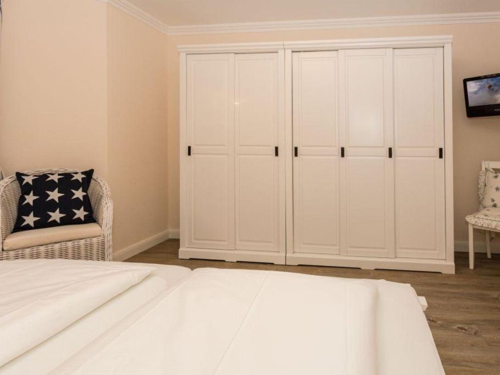 A bedroom with a bed, a white wardrobe, a wicker chair with a star-patterned pillow, and a television on the wall.