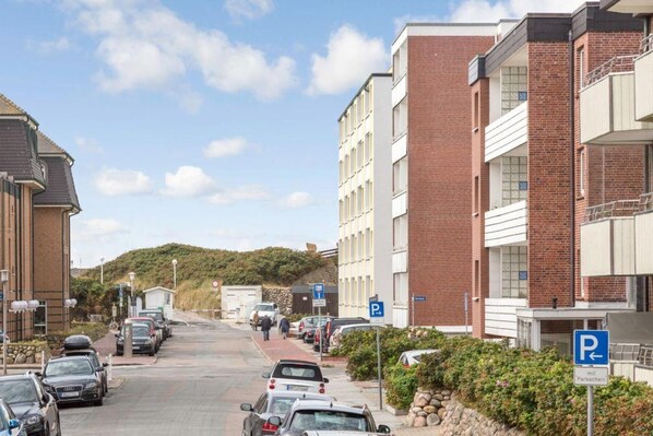 A residential street with parked cars, a clear sky, and a hilly landscape in the background. - Modern. Strandnah. Zentral. - Haus Margarethenstraße 2, Whg. 5 "juwel am Strand" - Objekt-id: Msw/02 (Sylt)