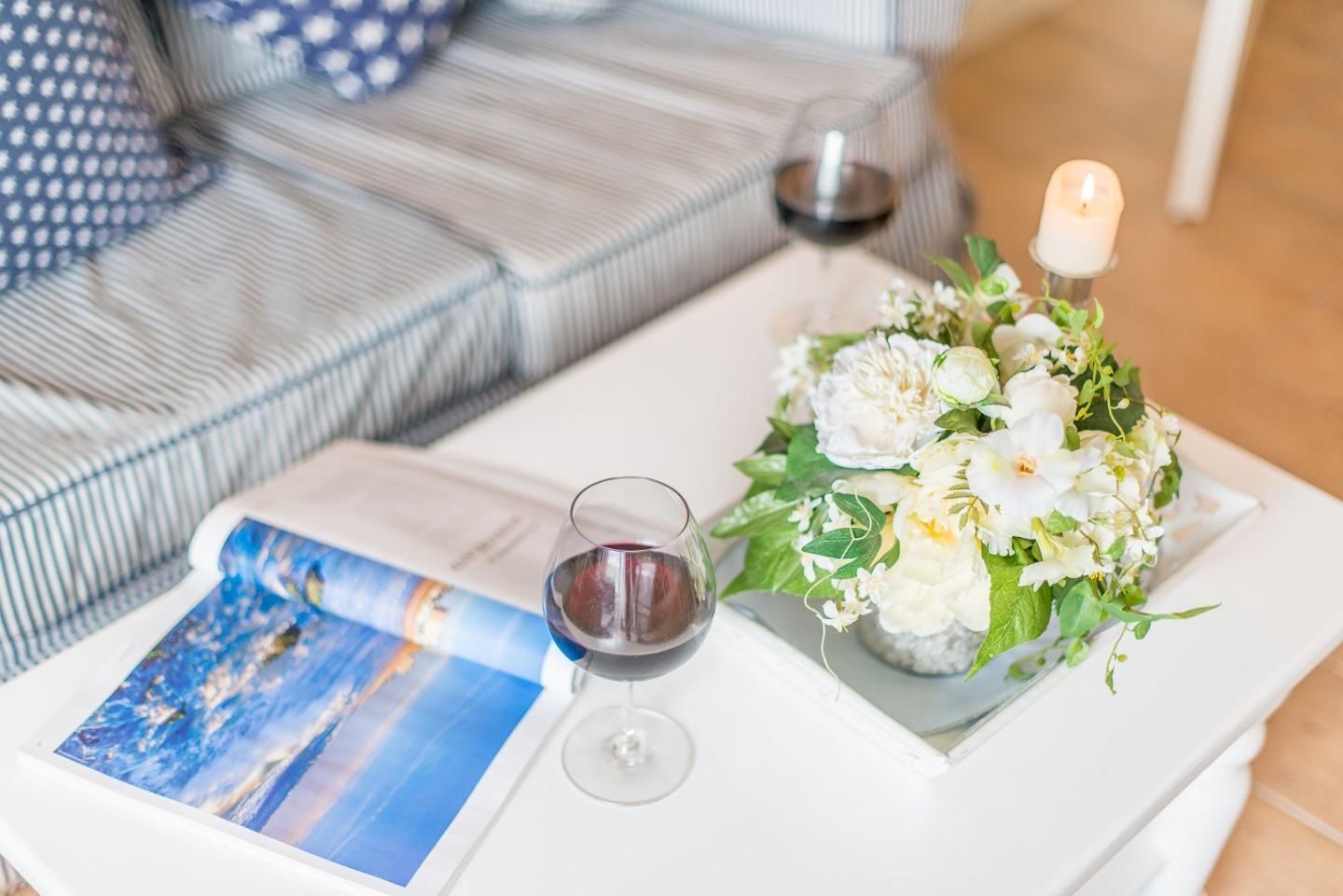 Una mesa blanca con una revista, dos copas de vino y un arreglo floral.
