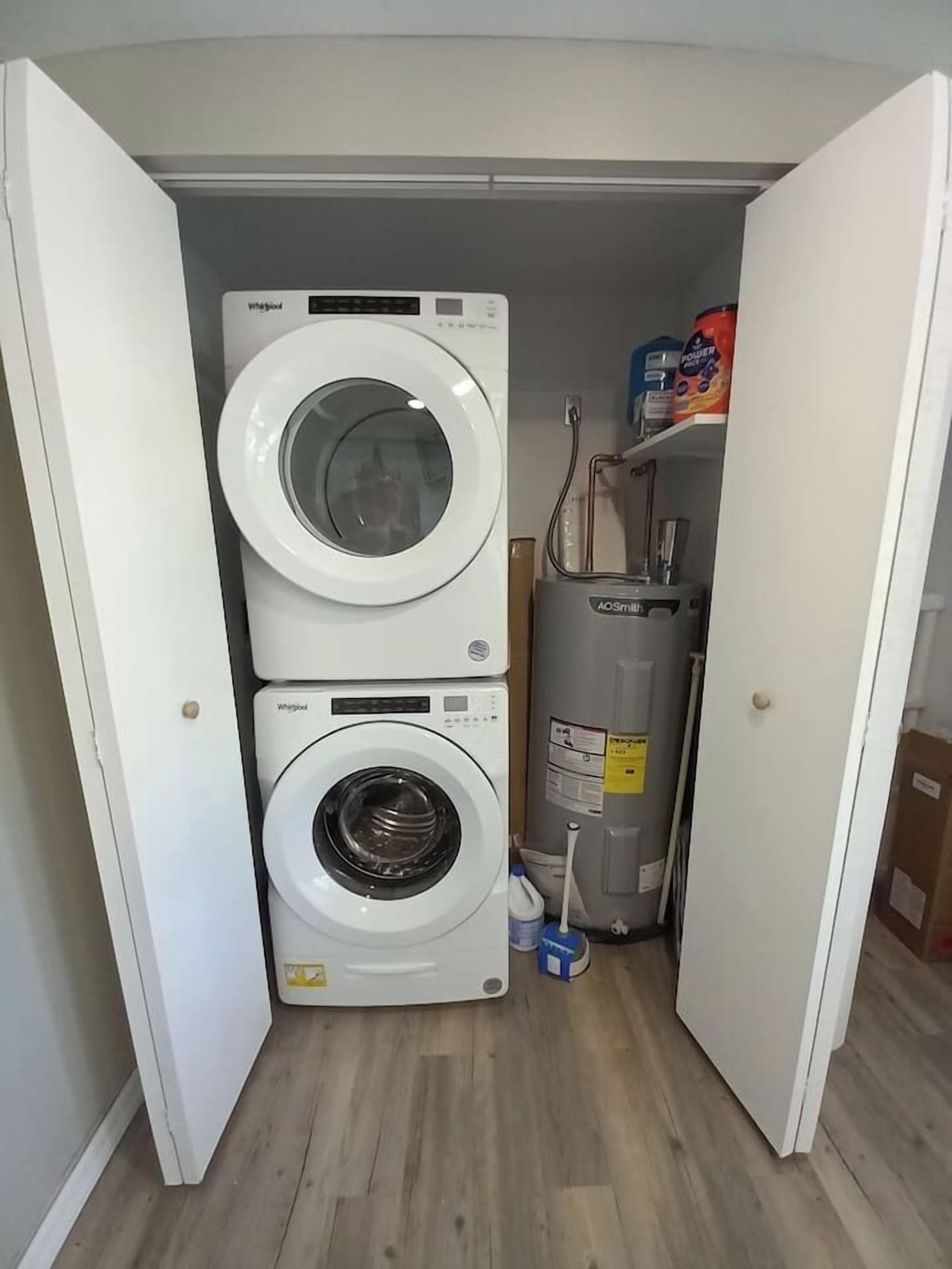 Une buanderie équipée de deux lave-linge Whirlpool superposés, d’un chauffe-eau et d’une étagère où sont rangées les lessives.