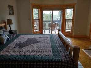Une chambre avec un grand lit, une tête de lit en bois, une vue sur une terrasse et des arbres par la fenêtre, et une lampe sur une table de chevet.