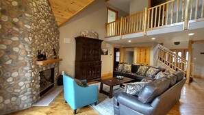 A spacious living area with a stone wall, a fireplace, a blue armchair, a leather sofa, and a wooden staircase.