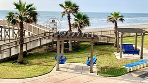 A wooden boardwalk leading to a beach with palm trees and a gazebo with benches. - Galveston  2/2 Gulf Front -  Heated Pool, 2 Hot Tubs, W/D in Unit (Galveston)
