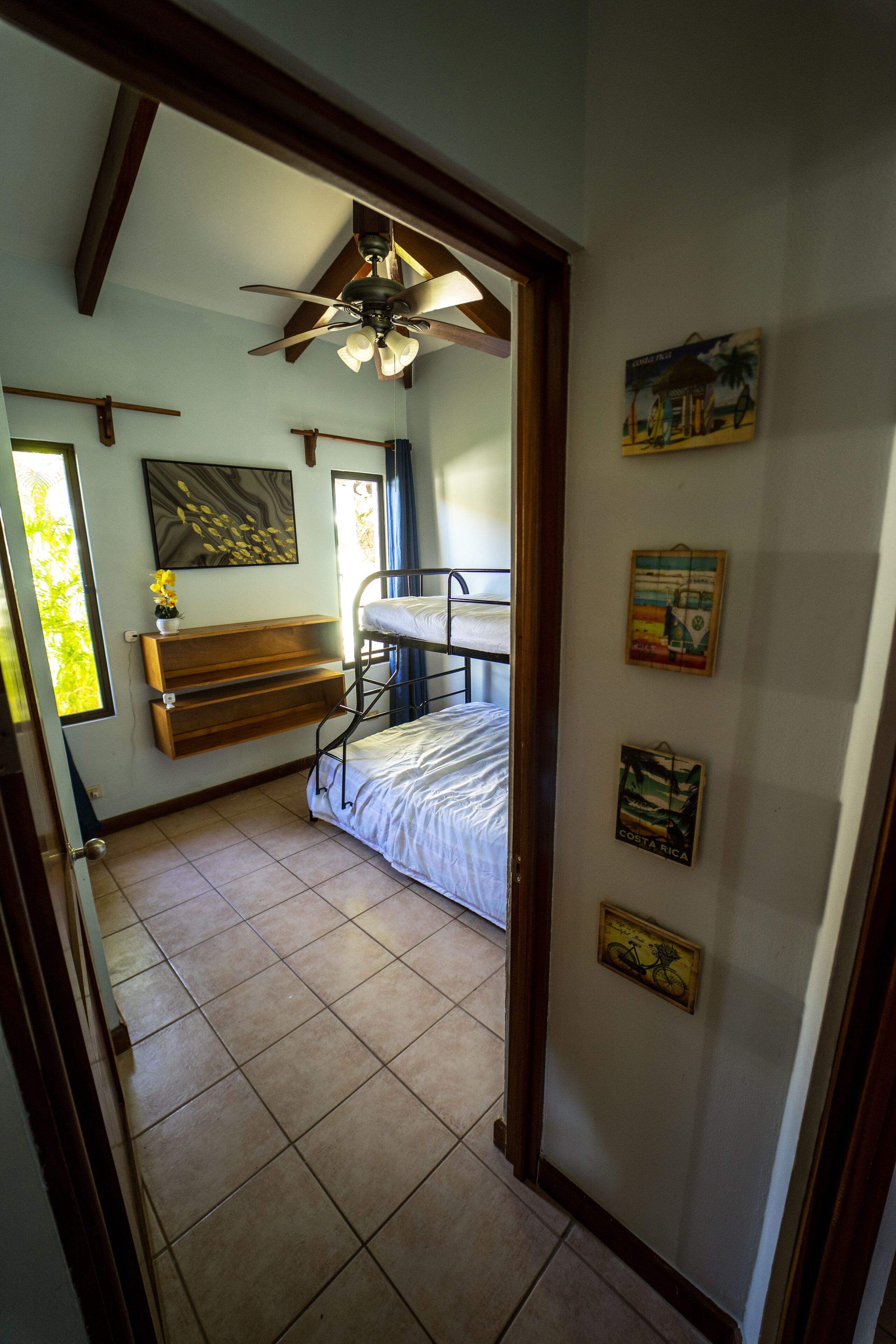 A room with a bunk bed, a ceiling fan, a window with curtains, a door, and wall decorations including a map of Costa Rica.