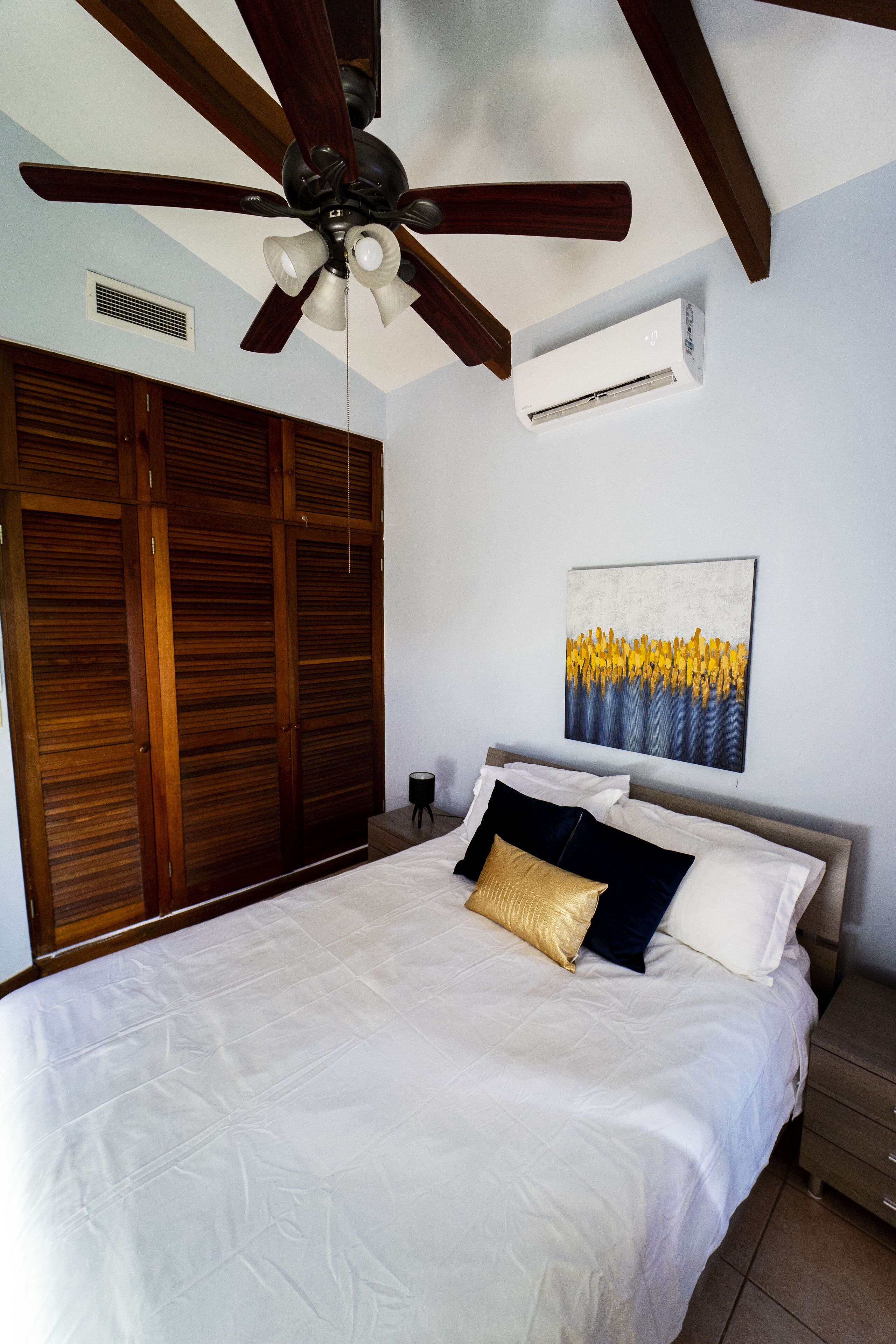 A bedroom with a bed, a ceiling fan, a wall-mounted air conditioner, wooden wardrobes, and a painting.
