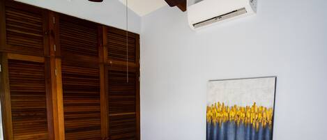 A bedroom with a bed, a ceiling fan, a wall-mounted air conditioner, wooden wardrobes, and a painting.