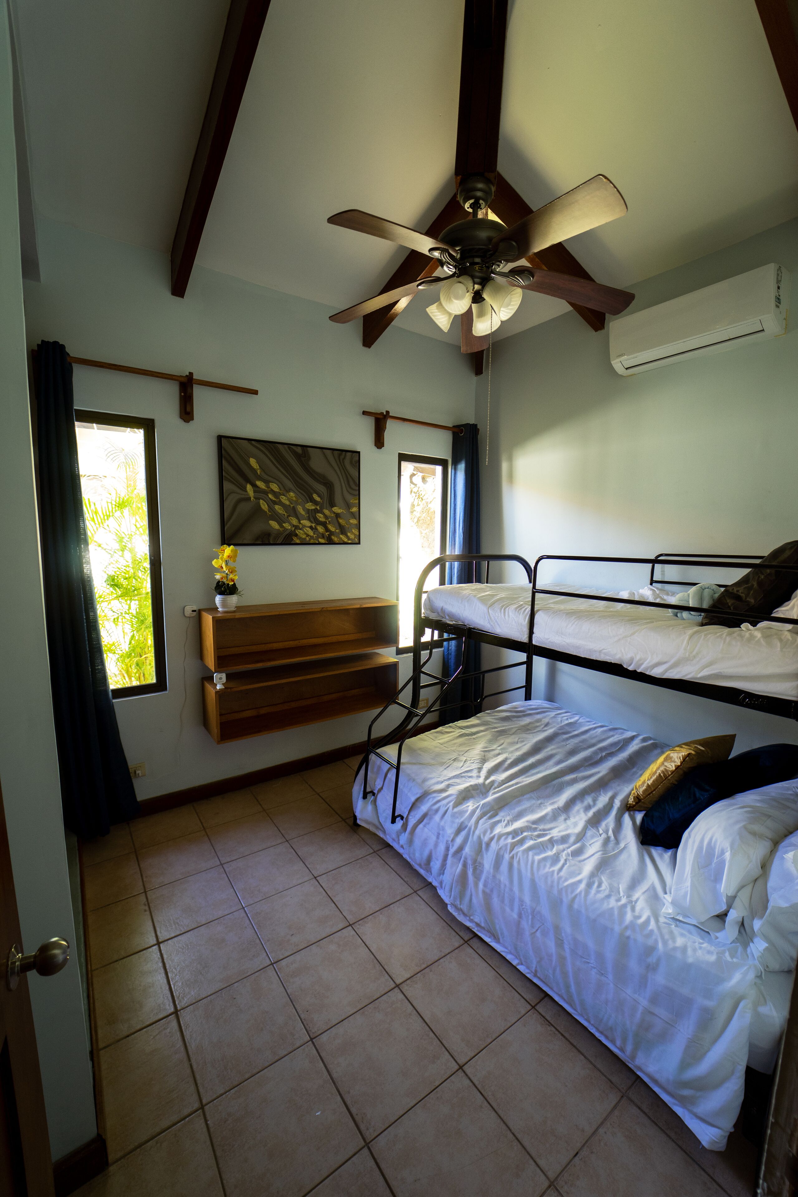 A room with two bunk beds, a ceiling fan, a window with curtains, a wall-mounted air conditioner, and a shelf with a plant.