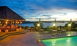 Ein Poolbereich mit Tischen und Stühlen, eine überdachte Lounge und Blick auf das Wasser.
