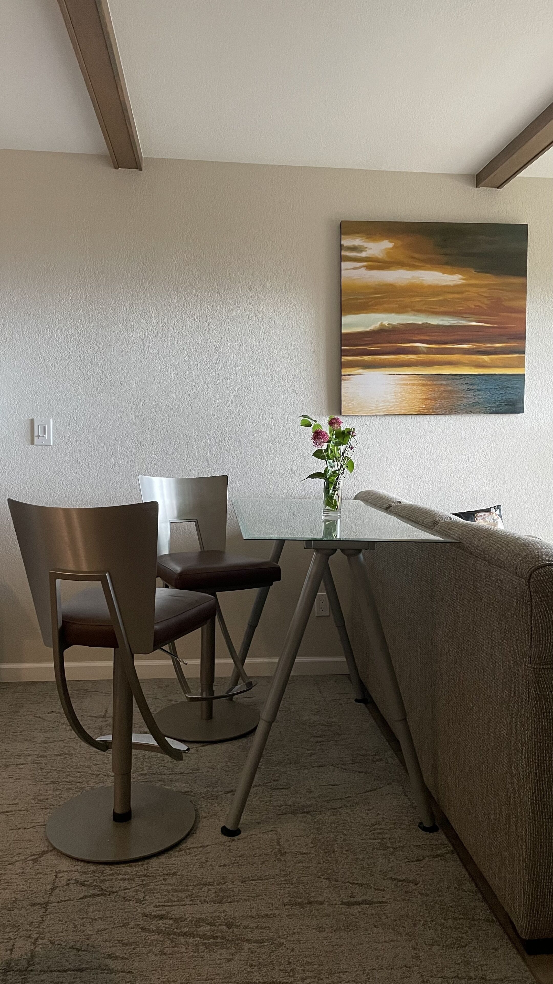 Ein modernes Wohnzimmer mit einem Esstisch mit Glasplatte, zwei Stühlen, einem Sofa und einem Sonnenuntergangsbild an der Wand.