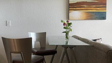 Ein modernes Wohnzimmer mit einem Esstisch mit Glasplatte, zwei Stühlen, einem Sofa und einem Sonnenuntergangsbild an der Wand.