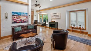 A spacious living room with a brown leather sofa, a coffee table, and a fireplace. There is a dining area with a table and chairs, and a large window with blinds. - Downtown Craftsman with King Bed and Private Yard - Walk Everywhere  (Bend)