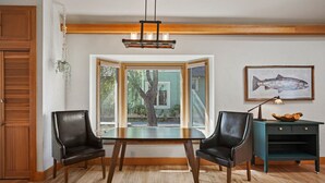 A dining area with a wooden table and two chairs, a hanging light fixture, a window with a view of a tree, and a fish painting on the wall. - Downtown Craftsman with King Bed and Private Yard - Walk Everywhere  (Bend)