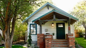 A small, single-story house with a green roof, a front porch with steps, and a brick chimney. - Downtown Craftsman with King Bed and Private Yard - Walk Everywhere  (Bend)