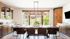 A modern kitchen with a dining table set for four, large windows, and a view of greenery.