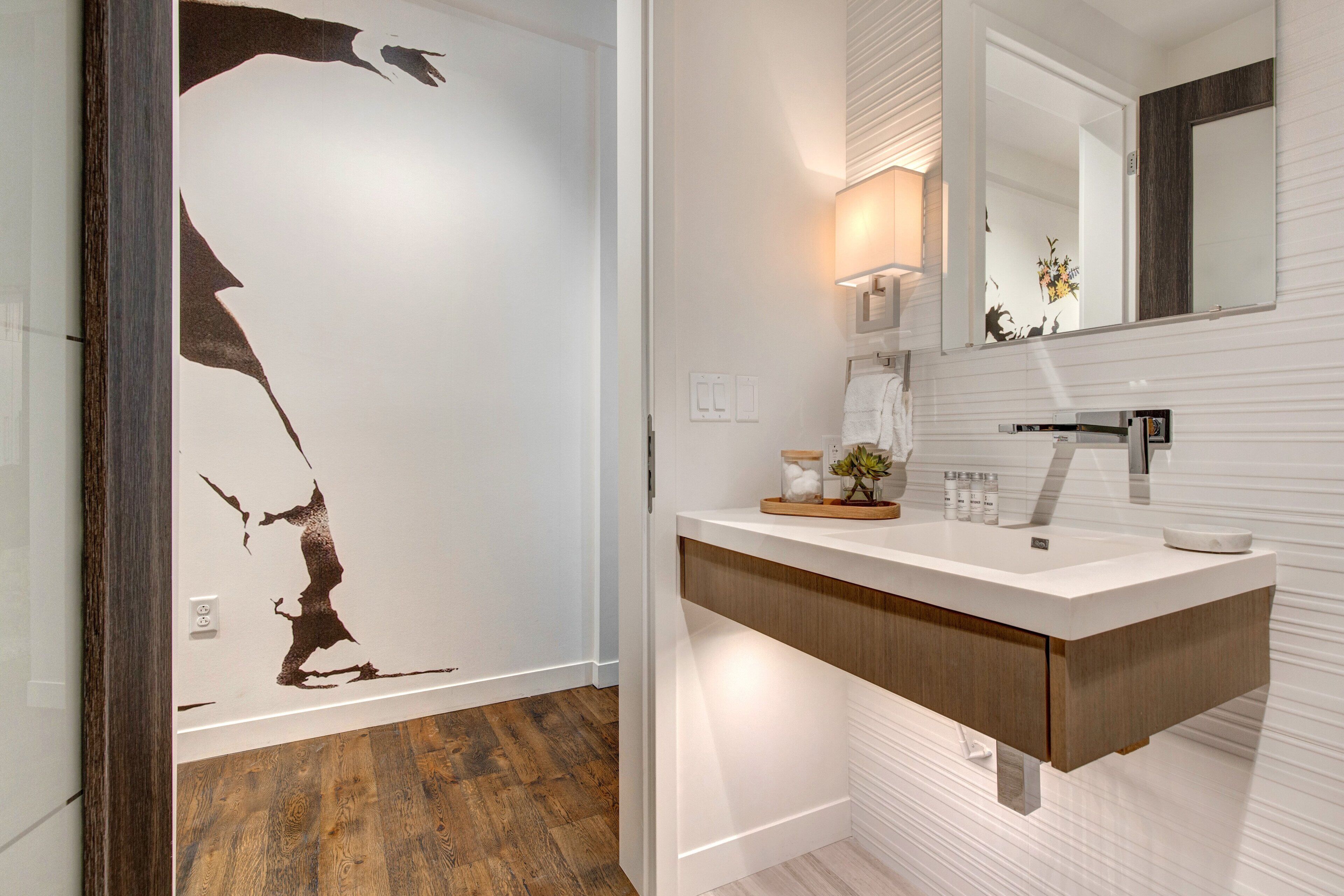 A bathroom with a wooden floor, a white sink with a wooden countertop, a mirror above the sink, and a wall with a large abstract artwork.
