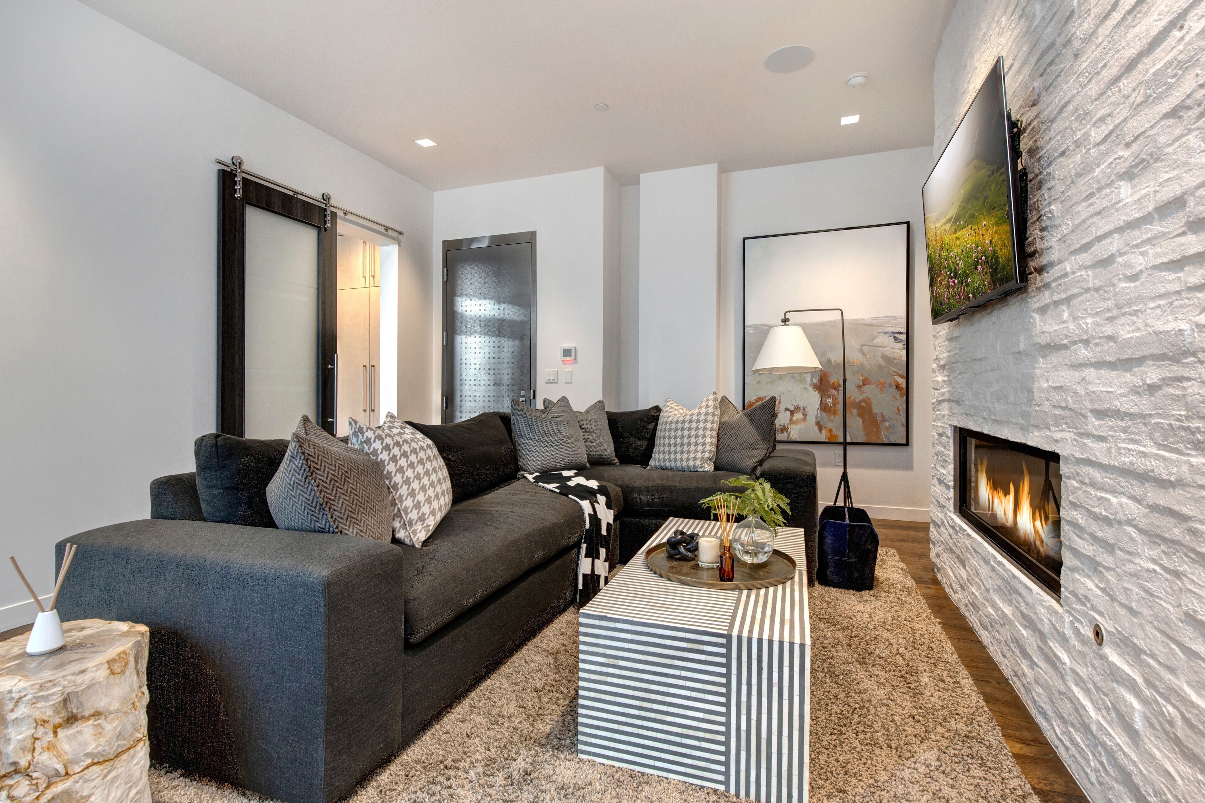 A modern living room with a fireplace, dark sofas, a striped coffee table, and a large abstract painting on the wall.