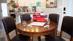 A dining table with four chairs, a board game, and two glasses of wine in a modern kitchen with white cabinets and a farmhouse-style painting.