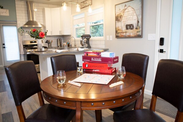 A dining table with four chairs, a board game, and two glasses of wine in a modern kitchen with white cabinets and a farmhouse-style painting. - Cozy Bungalow w/ Hot Tub & Fire Pit, Walk Downtown (Loveland)