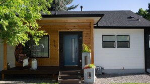 A modern two-story house with a wooden front porch, a blue door, and a small garden area with potted plants.