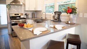 A modern kitchen with white cabinets, a stainless steel range hood, a large island with breakfast items, and a window with a view of greenery.