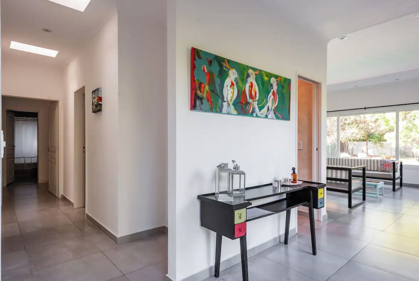 A hallway with a modern desk, a colorful abstract painting, and a view of the outdoors through large windows.
