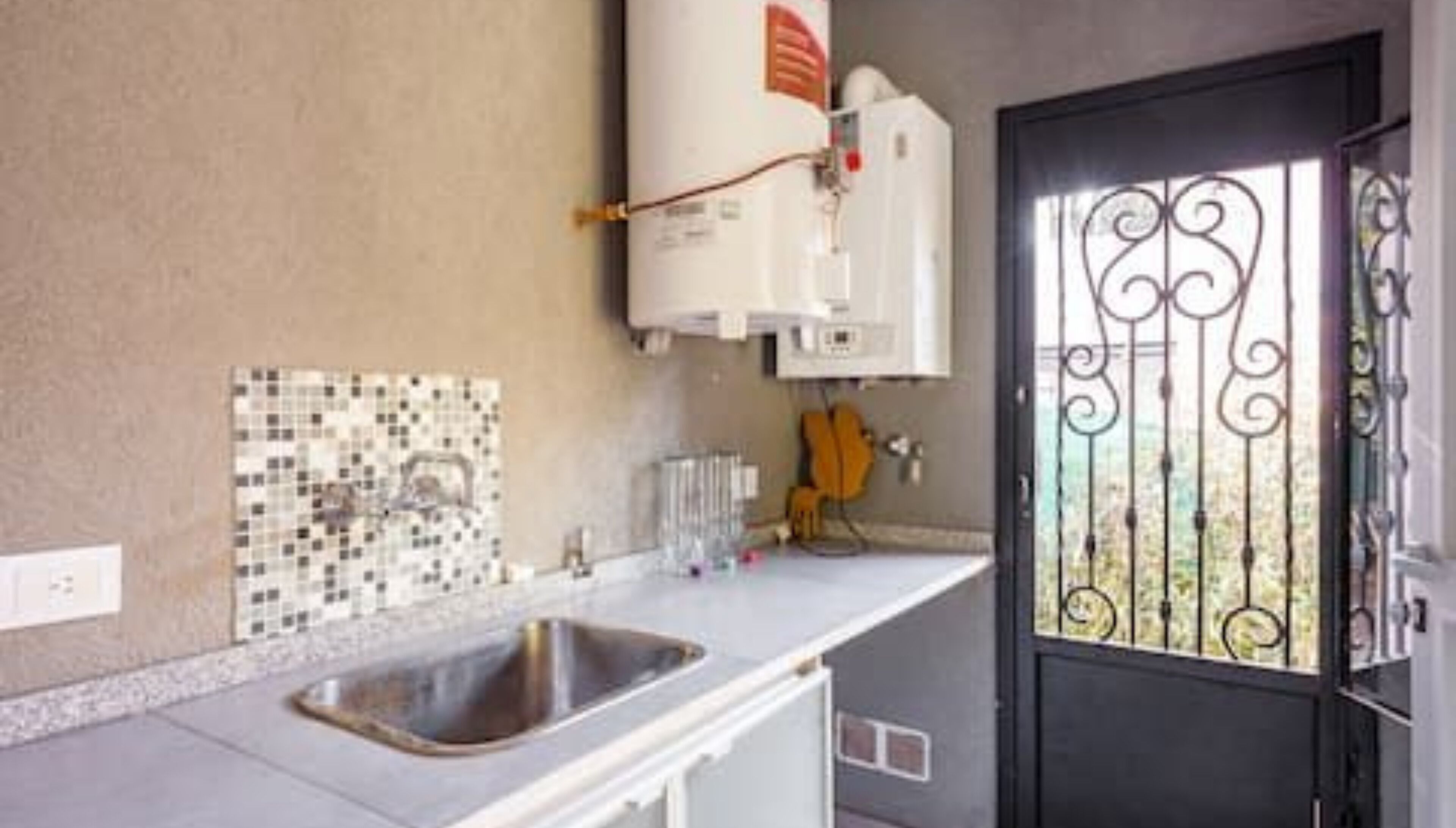 Una cocina con un lavamanos, un calentador de agua blanco, salpicadero de azulejos y una puerta negra con reja metálica.