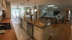 A spacious kitchen with granite countertops, white cabinetry, and a central island. There is a dining area with a table and chairs, and a living room with a sofa and a TV. - OCEAN BREEZE IS A LUXURY GETAWAY THAT HAS IT ALL (Ormond Beach)