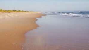 A wide sandy beach with gentle waves, a clear sky, and distant power lines. - OCEAN BREEZE IS A LUXURY GETAWAY THAT HAS IT ALL (Ormond Beach)