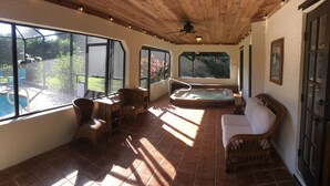 A sunlit patio area with a hot tub, wicker furniture, and large windows overlooking a garden. - OCEAN BREEZE IS A LUXURY GETAWAY THAT HAS IT ALL (Ormond Beach)