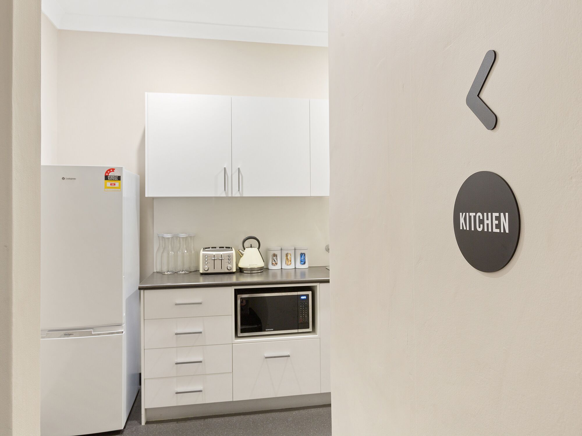 A modern kitchen with white cabinets, a microwave, a toaster, and a kettle. There is a 'KITCHEN' sign on the wall.
