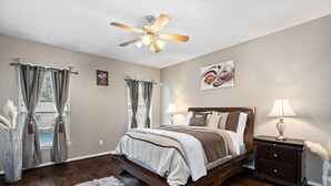 A bedroom with a wooden bed, two bedside tables with lamps, a ceiling fan, and a large window with curtains.