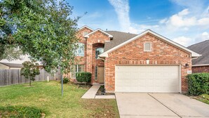 A two-story brick house with a white garage door, a front yard with grass and a small tree, and a clear blue sky.