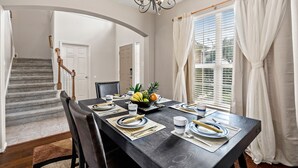 A dining area with a dark wooden table set for four, a chandelier, and a window with white curtains.
