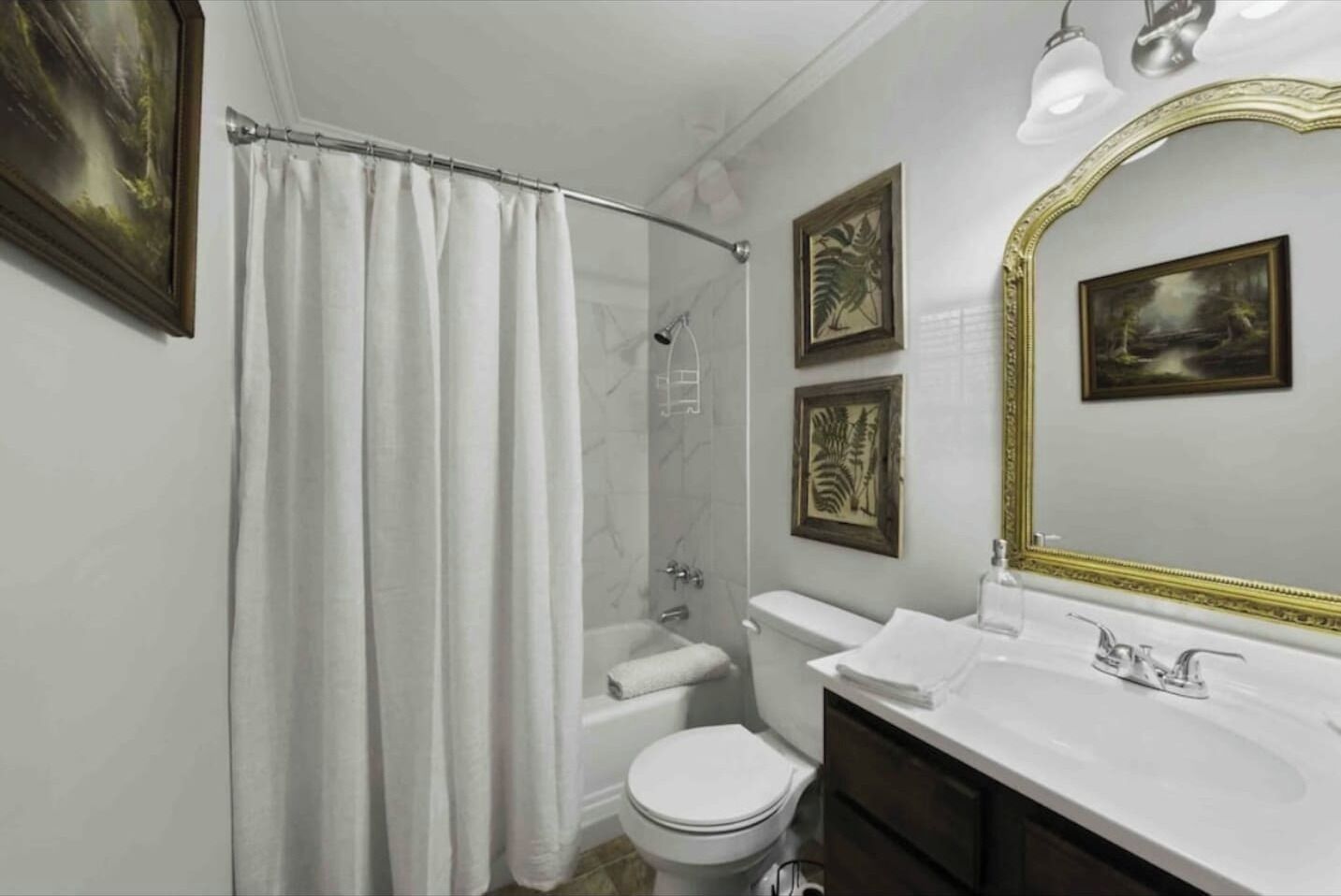 Une salle de bain avec une douche, des toilettes, un lavabo et un grand miroir avec un cadre doré. Des reproductions encadrées de motifs botaniques ornent le mur.