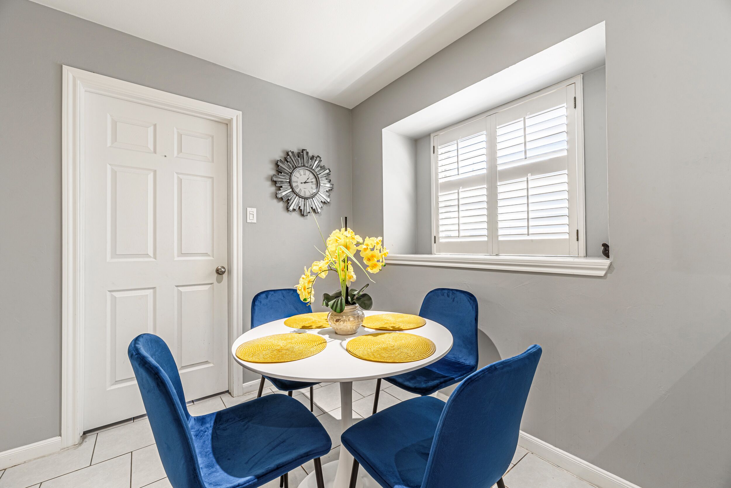 Un coin repas avec une table ronde, des chaises bleues, des sets de table jaunes et un vase rempli de fleurs jaunes.