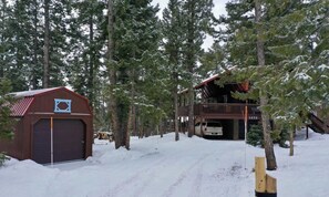 En stuga med ett rött tak och ett garage, omgiven av höga tallar och täckt av snö.