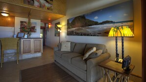 Een woonkamer met een bank, een tafel met een lamp, een schilderij van een strand en een keuken met een koelkast en een plant.