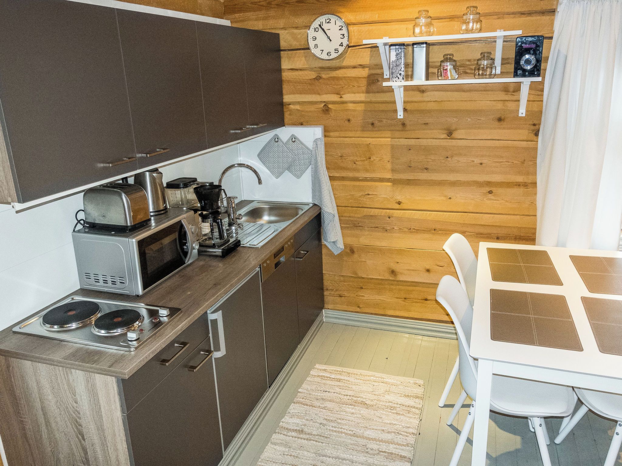 A compact kitchen with a microwave, coffee maker, and stove. There's a wooden wall, a clock, and a shelf with jars.