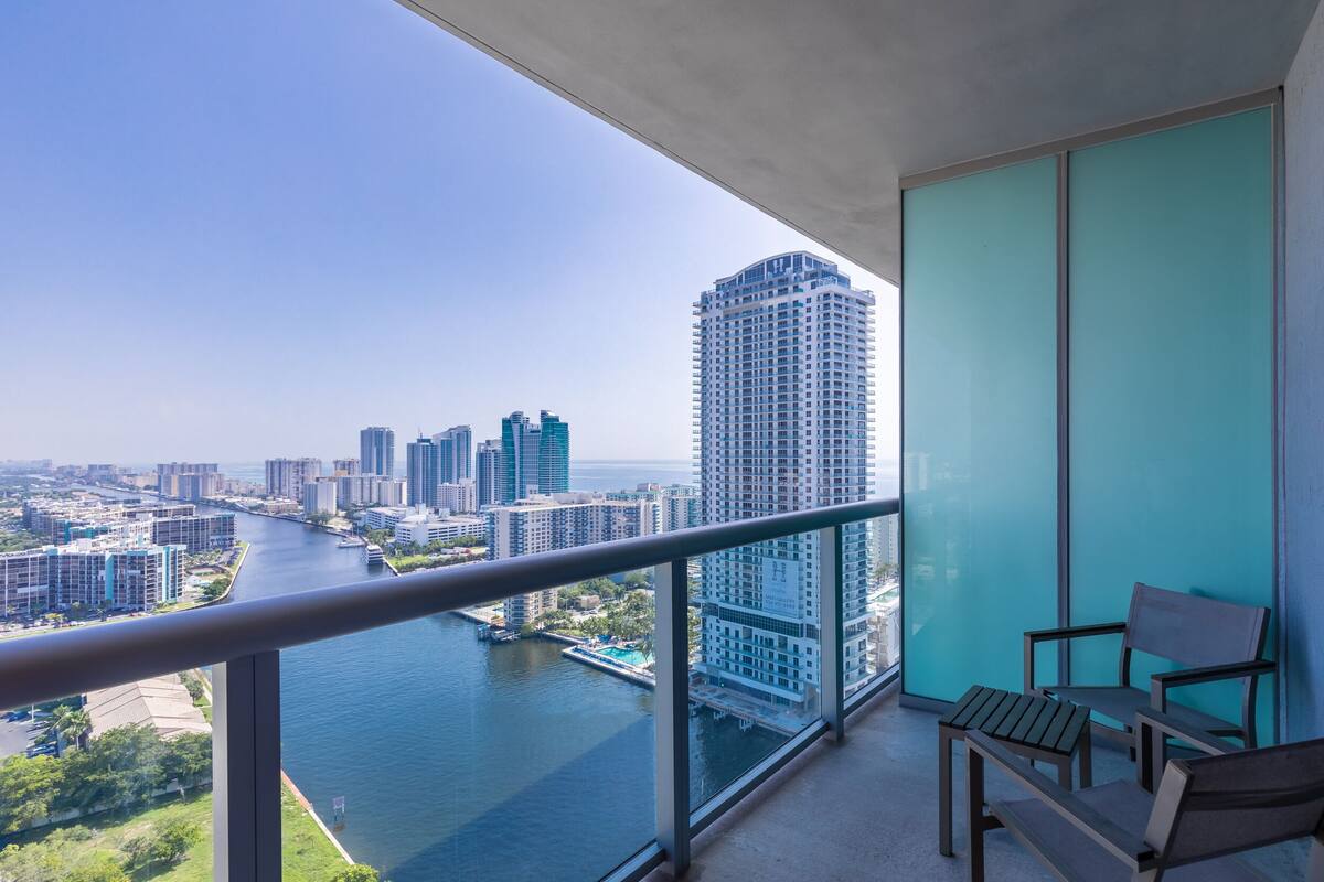 Balcón con vista a un río y al paisaje urbano, con un divisor de vidrio y dos sillas de madera.